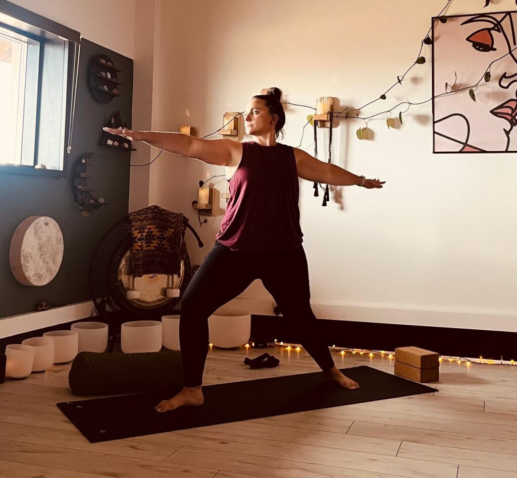 KynWellbeing practitioner in a maroon sleeveless top and black leggings performing a Warrior II yoga pose in a sunlit studio with candles and artwork.